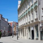 Eine ruhige, sonnige Straße gesäumt von historischen europäischen Gebäuden und einer großen Topfpflanze im Vordergrund.