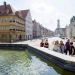 Touristengruppe neben einem Brunnen in einer europäischen Stadtstraße mit historischen Gebäuden und einem Uhrturm im Hintergrund.