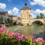 Malerische Aussicht auf ein historisches Gebäude auf einer Brücke über einen Fluss, umgeben von Blumen und traditionellen Häusern, unter einem blauen Himmel.