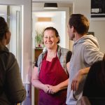 Eine lächelnde Frau in einer Schürze begrüßt die Kunden am Eingang eines gemütlichen Cafés.