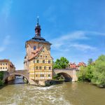 Historisches Gebäude mit einem Turm auf einer Brücke über einem Fluss, umgeben von Bäumen und traditioneller Architektur.