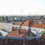 Blick von der Dachterrasse auf eine Stadtlandschaft mit roten Ziegeldächern und einem Fernsehturm in der Ferne unter einem klaren Himmel.