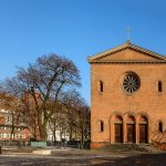 Rote Backsteinkirche mit Rosettenfenster und Kreuz, umgeben von kahlen Bäumen und einem klaren blauen Himmel.