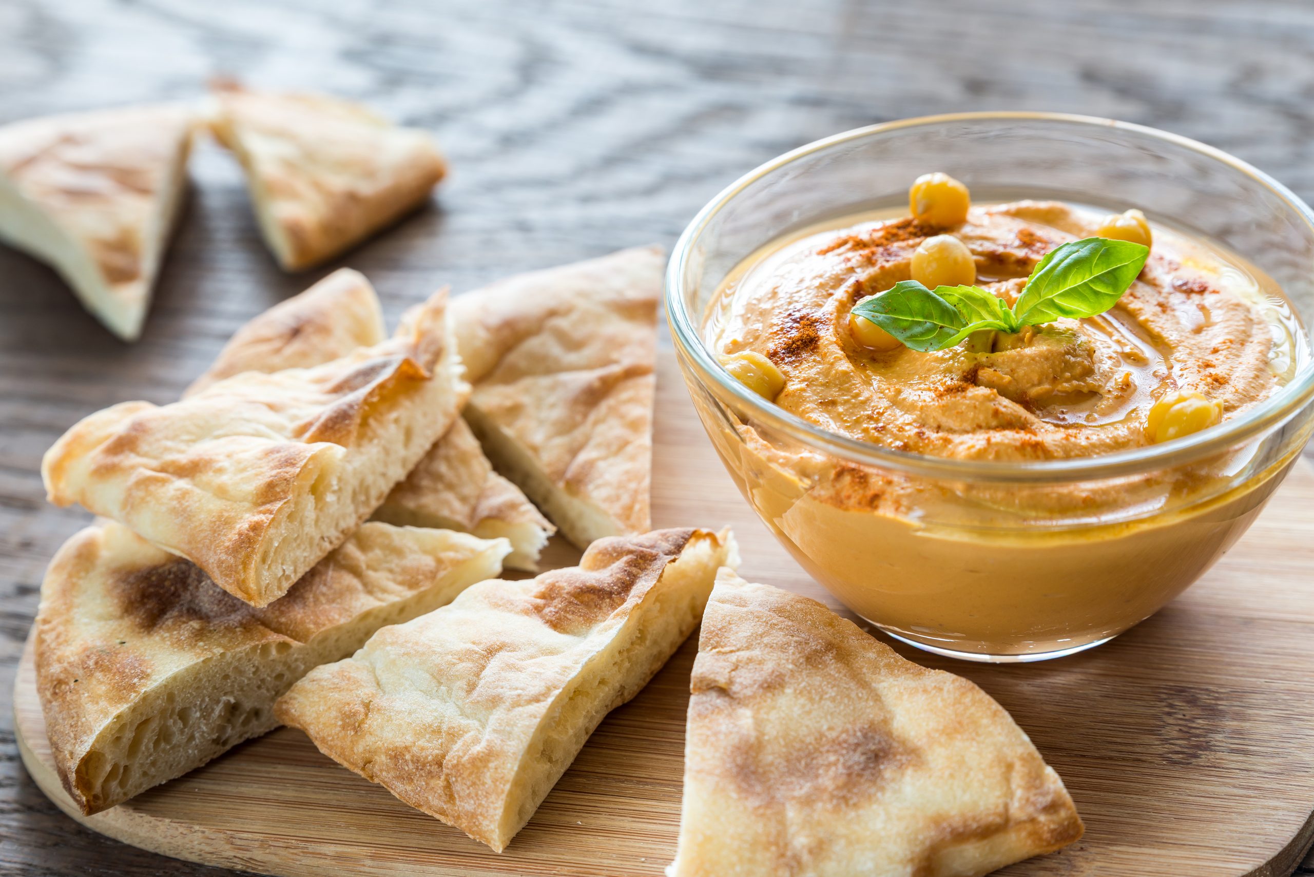 Eine köstliche Schüssel Hummus, garniert mit Kichererbsen und Basilikum, perfekt für einen Betriebsausflug, begleitet von geschnittenem Fladenbrot, elegant angerichtet auf einem Holzbrett.