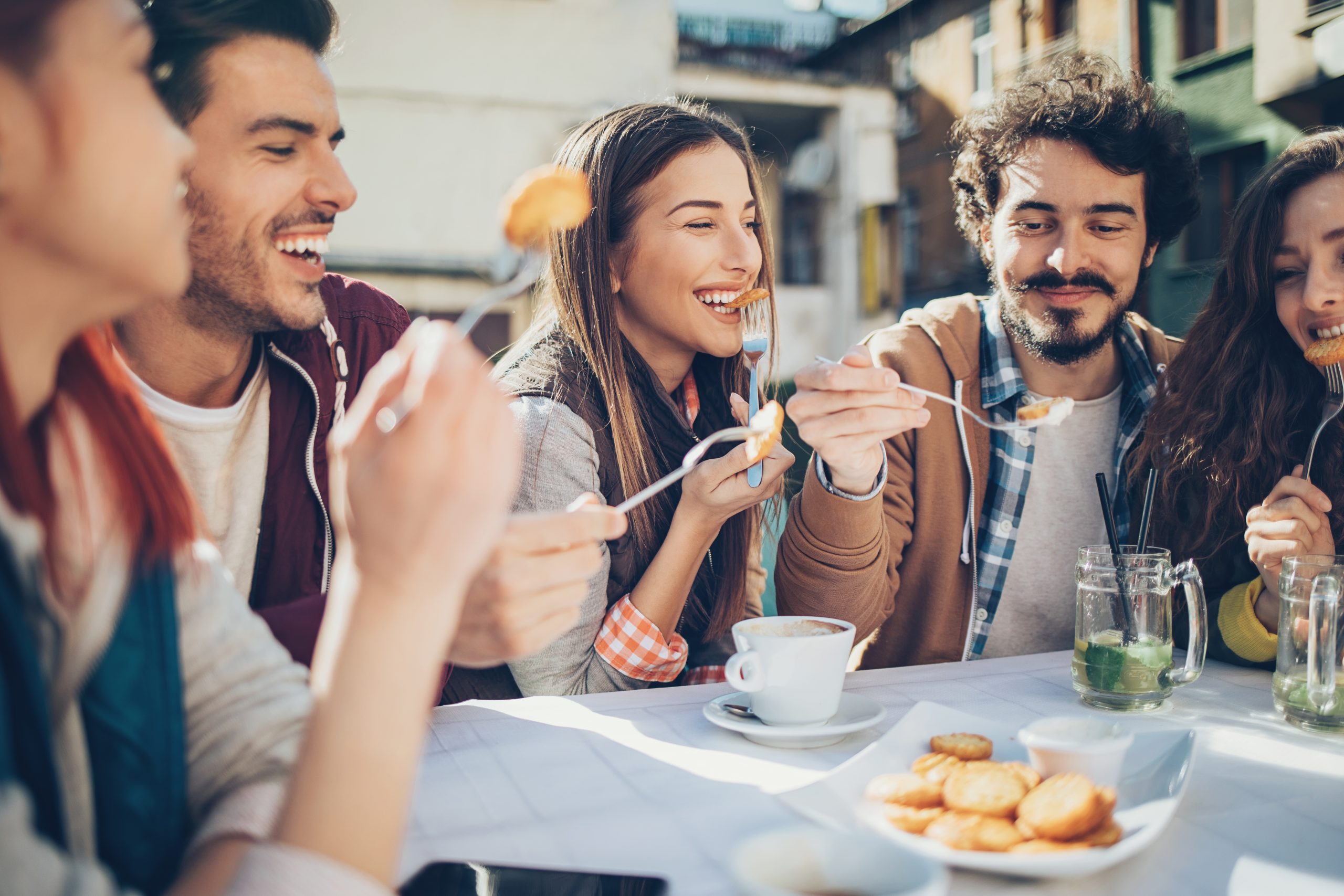 Eine Gruppe von Freunden, möglicherweise auf einem Betriebsausflug, genießt Snacks und Getränke in einem Straßencafé, lächelt und führt angeregte Gespräche.