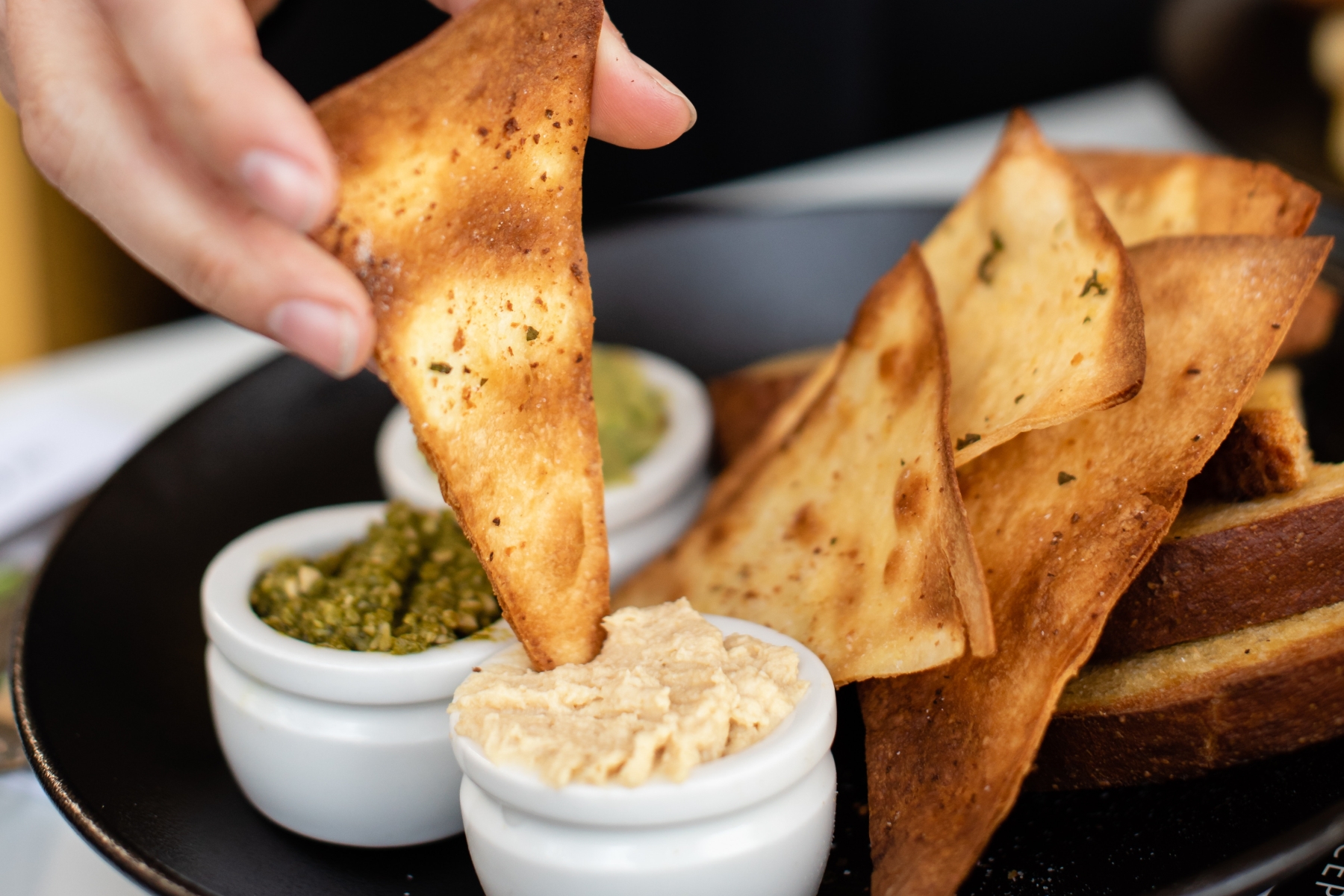 Während eines Betriebsausflugs genießt jemand das einfache Vergnügen, einen gewürzten Pita-Chip in Hummus zu tunken, während in einer kleinen Schüssel Pesto bereitsteht.