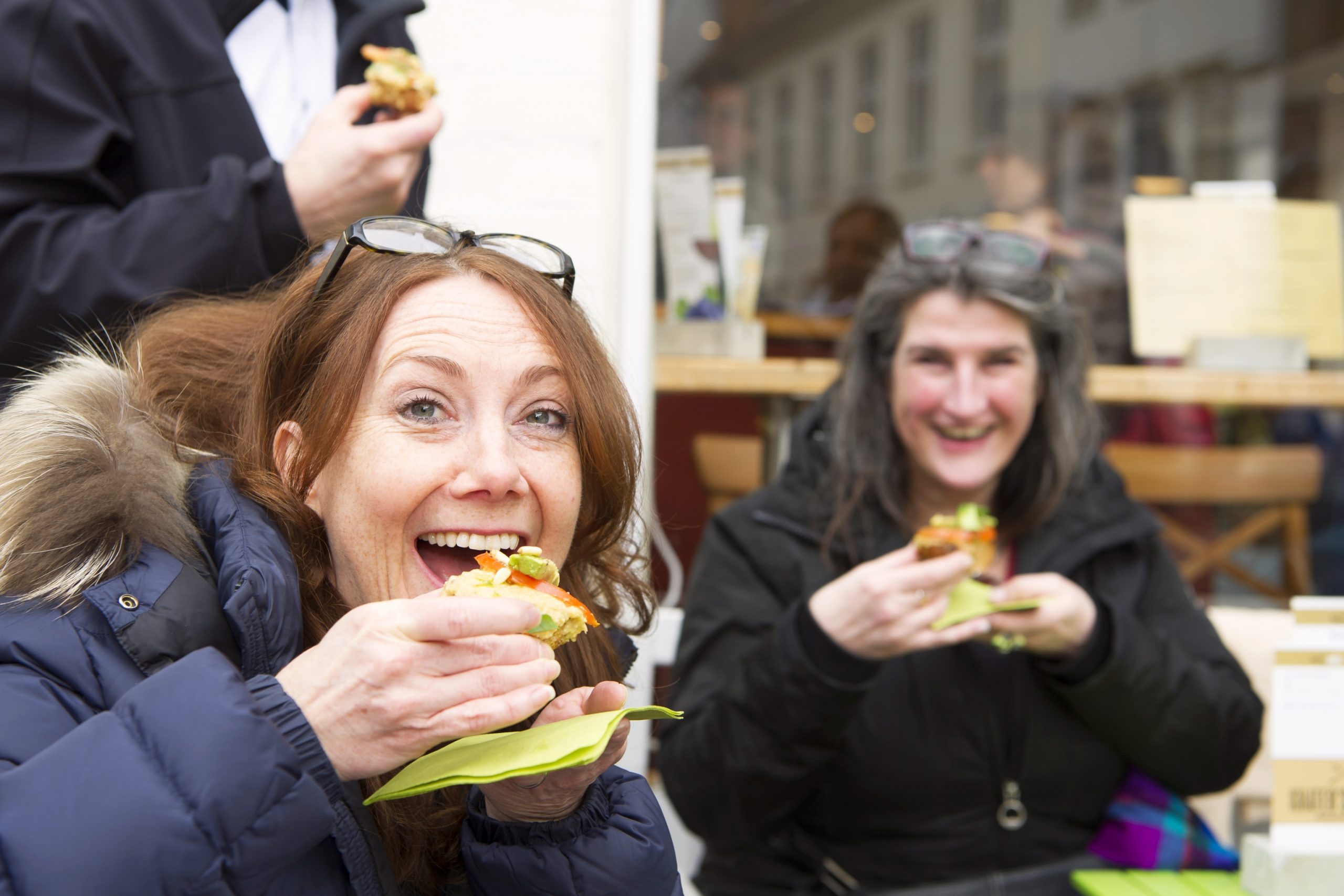 Zwei Frauen genießen einen sonnigen Betriebsausflug und lächeln, während sie im Freien Sandwiches essen, wobei eine direkt in die Kamera blickt.