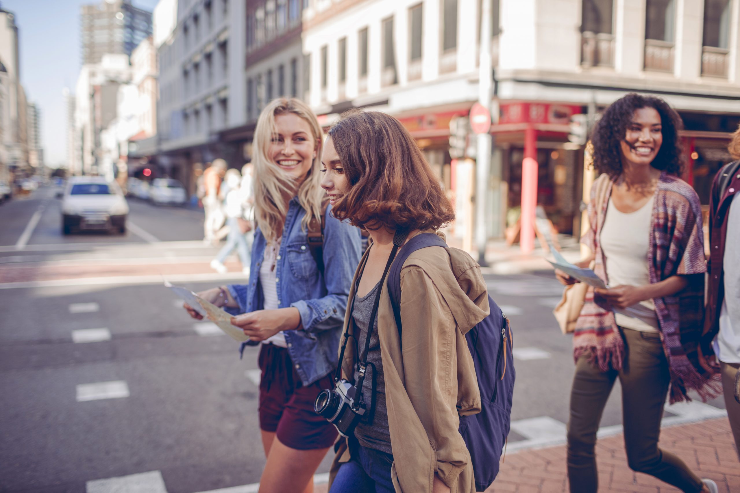 Vier Freunde überqueren auf einem Betriebsausflug lächelnd und mit Karten in den Händen eine belebte Stadtstraße, im Hintergrund sind hoch aufragende Gebäude zu sehen.