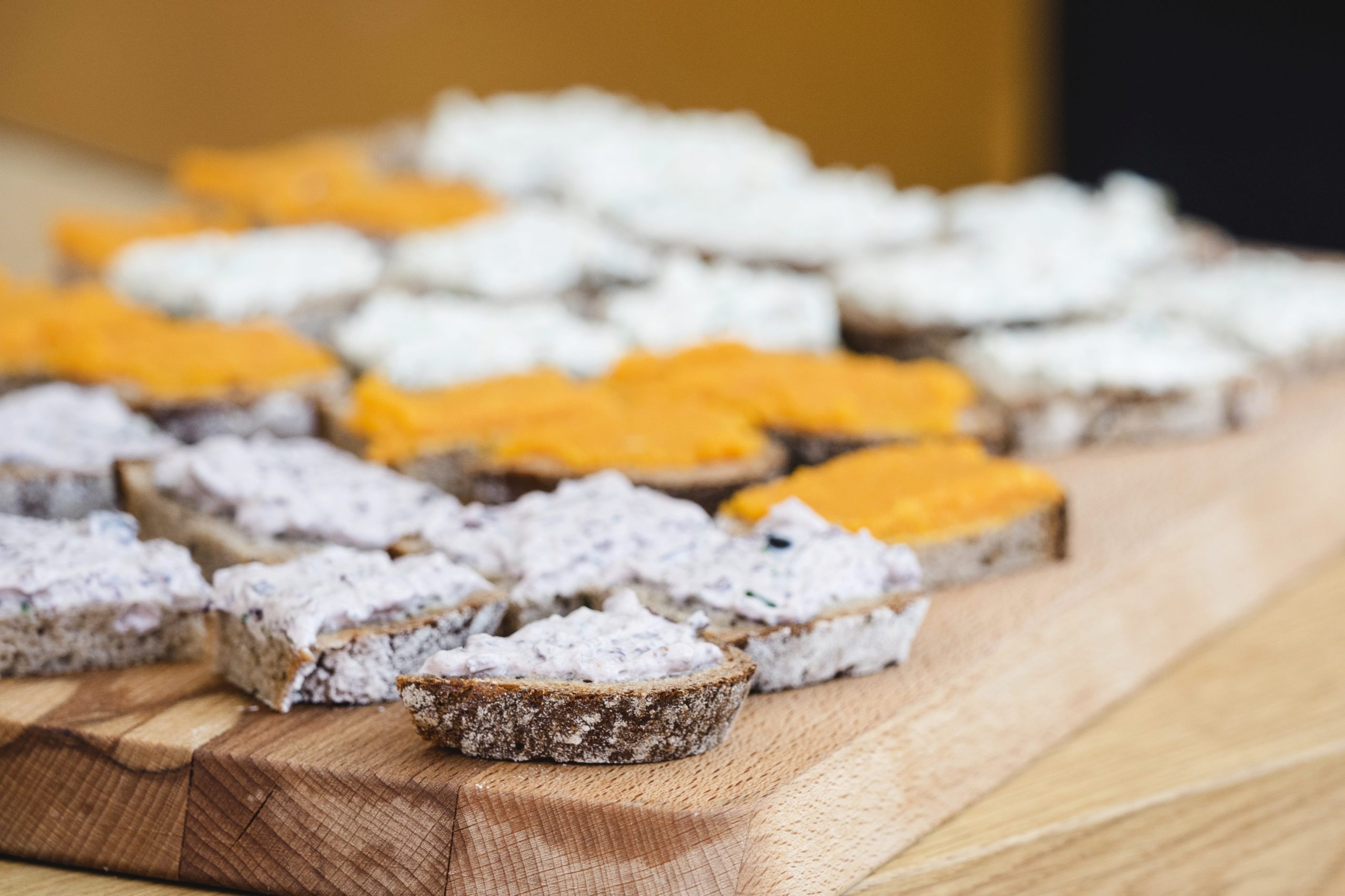 Auf einem Holzbrett angerichtete Brotscheiben mit verschiedenen Aufstrichen, unter anderem cremigem Orangen- und Weißbrot – perfekt für ein gemütliches Betriebsausflug-Treffen.