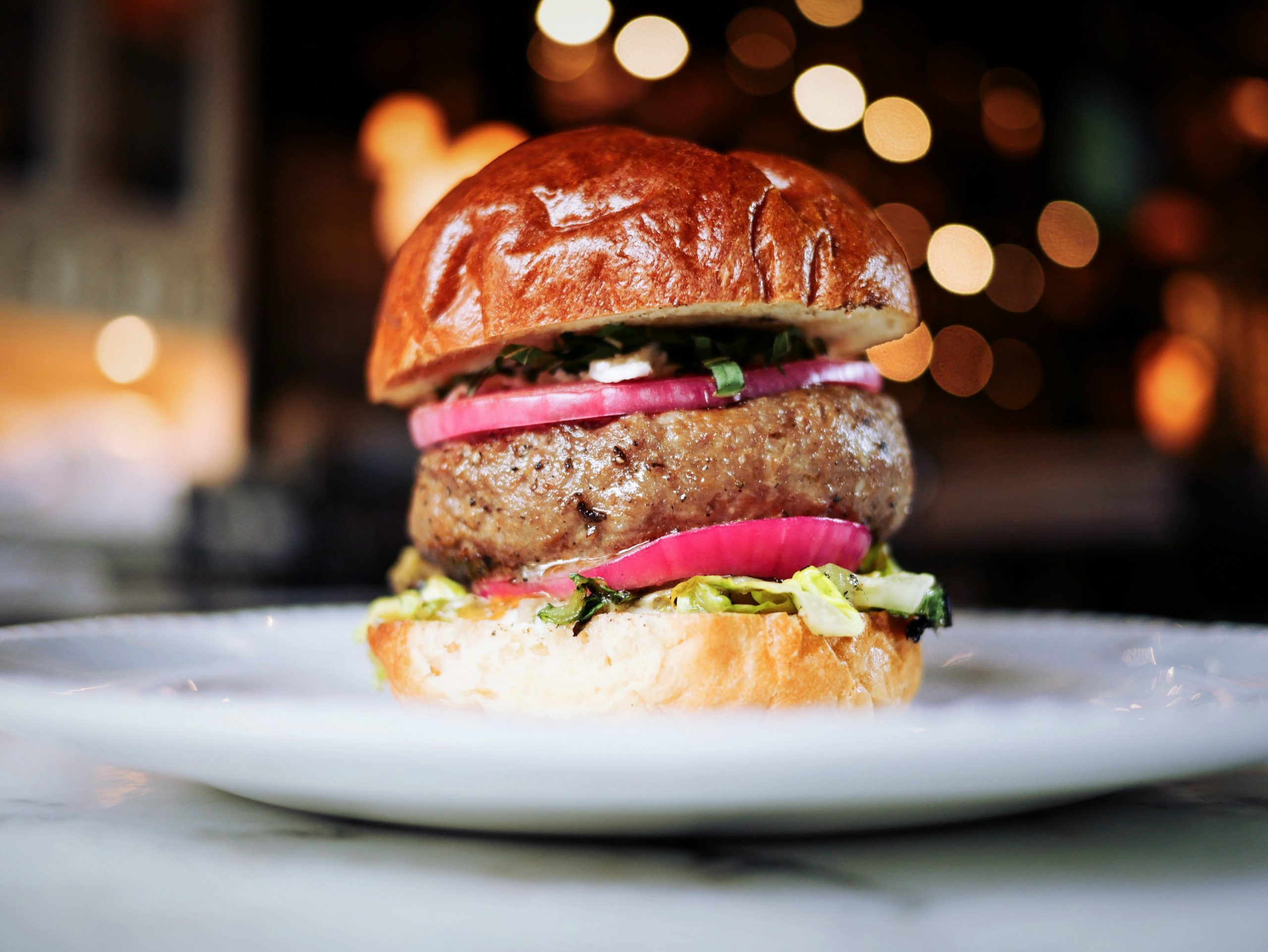 Eine Nahaufnahme eines Gourmet-Burgers, perfekt für ein Betriebsausflug-Festmahl, mit einem Rindfleisch-Patty, frischem Salat und würzigen eingelegten Zwiebeln auf einem glänzenden Brötchen, alles elegant präsentiert auf einem weißen Teller.