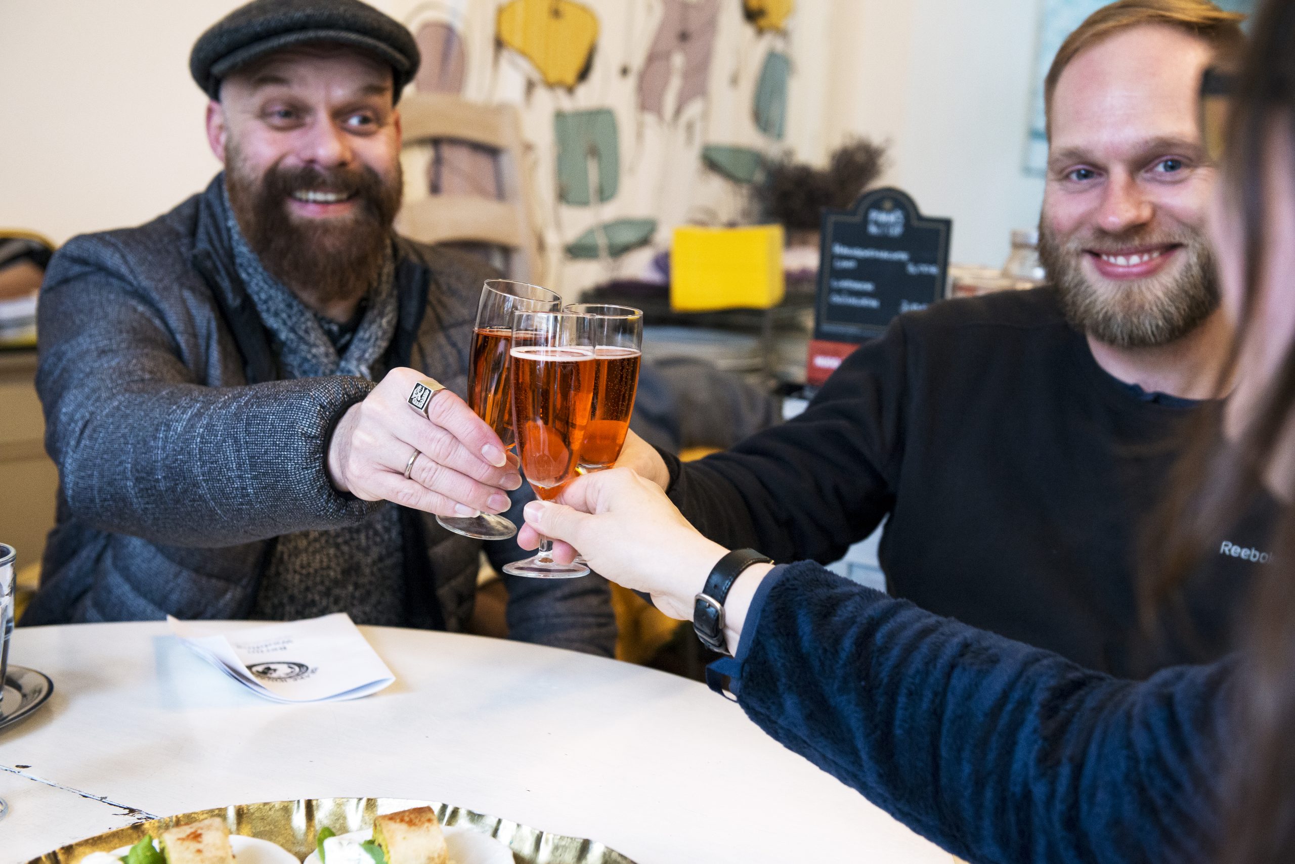 Drei Kollegen stoßen in gemütlicher Atmosphäre an einem mit Essen gedeckten Tisch an und feiern lächelnd ihren erfolgreichen Betriebsausflug.