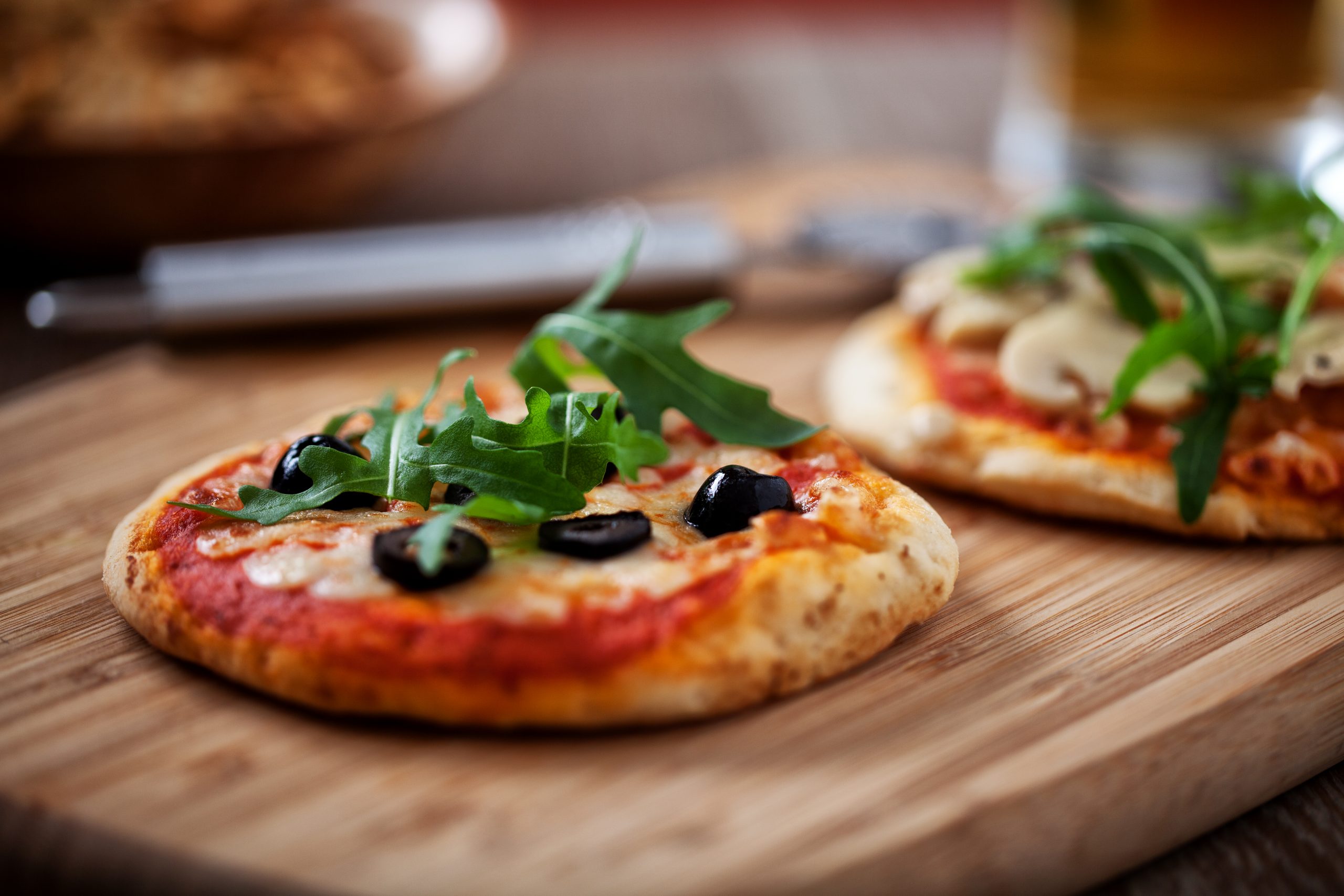 Zwei Minipizzas mit Rucola, Oliven und Käse liegen einladend auf einem Holzbrett und eignen sich perfekt zum Teilen bei einem entspannten Betriebsausflug. Der unscharfe Hintergrund trägt zur gemütlichen Atmosphäre bei.