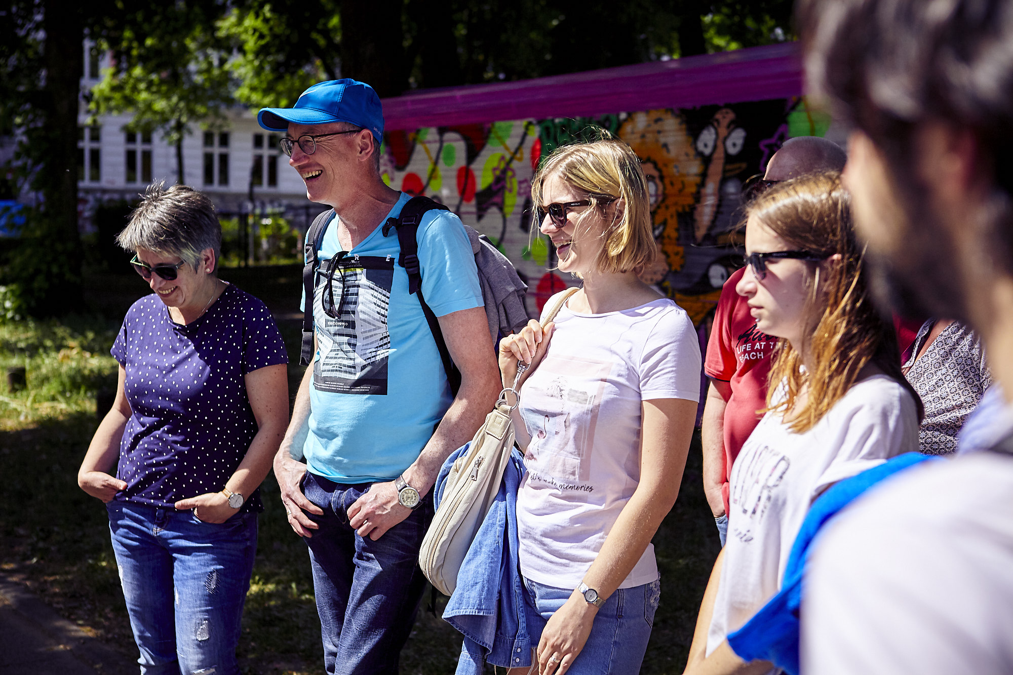 Eine Gruppe von Menschen steht auf einem lebhaften Betriebsausflug im Freien, lächelt und trägt Freizeitkleidung, während Bäume und Graffiti die lebendige Kulisse bemalen.