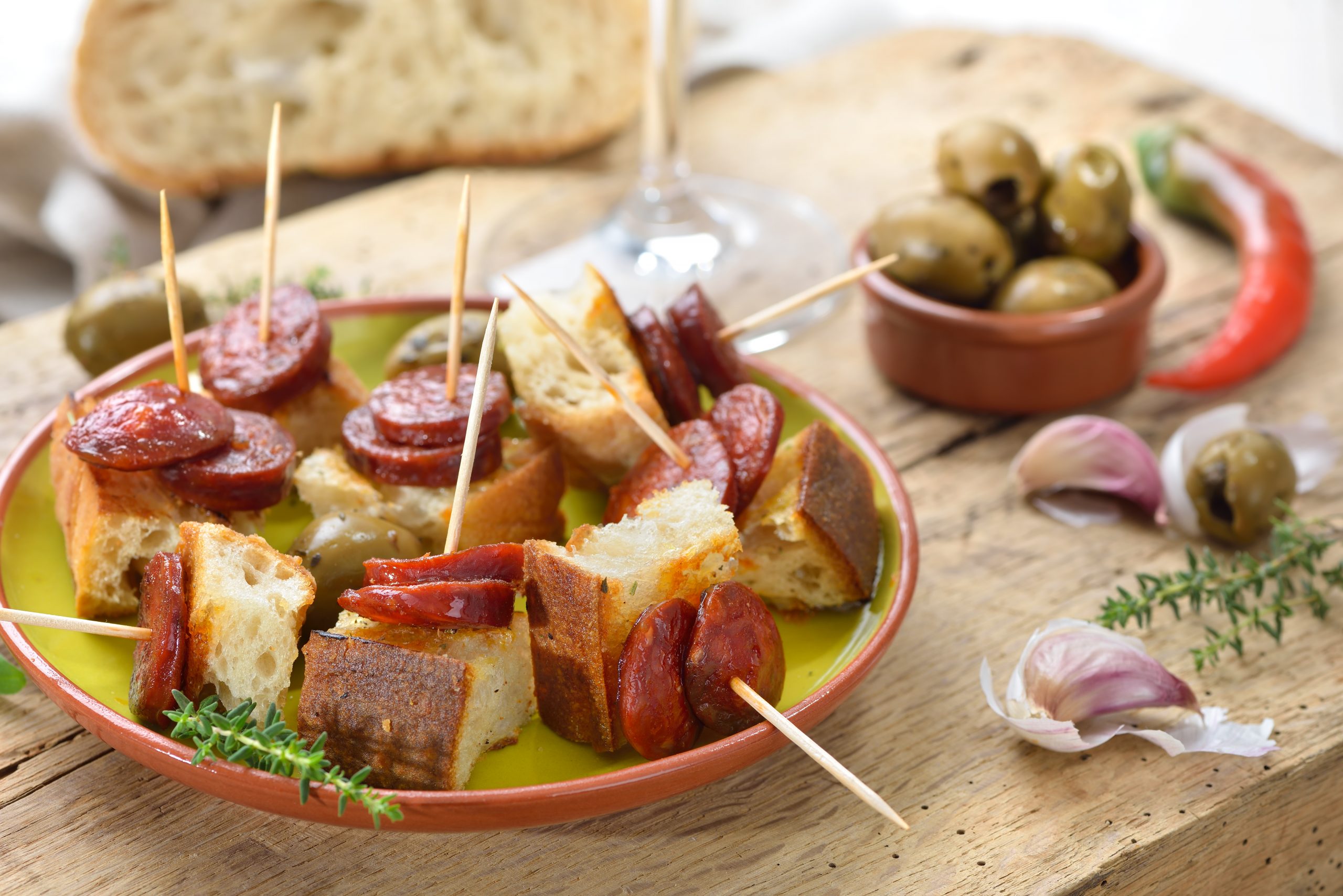 Ein Teller mit Chorizo, Brot, Oliven und Knoblauch mit Zahnstochern steht einladend auf einem Holztisch, akzentuiert mit roten Chilis und Kräutern – ein perfektes Arrangement für ein Betriebsausflug-Picknick.