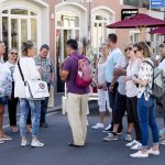 Eine Gruppe von Menschen steht auf einer Straße, manche mit Rucksäcken, plaudert angeregt über ihren Betriebsausflug und genießt die lebhafte Atmosphäre und die Gesellschaft der anderen.