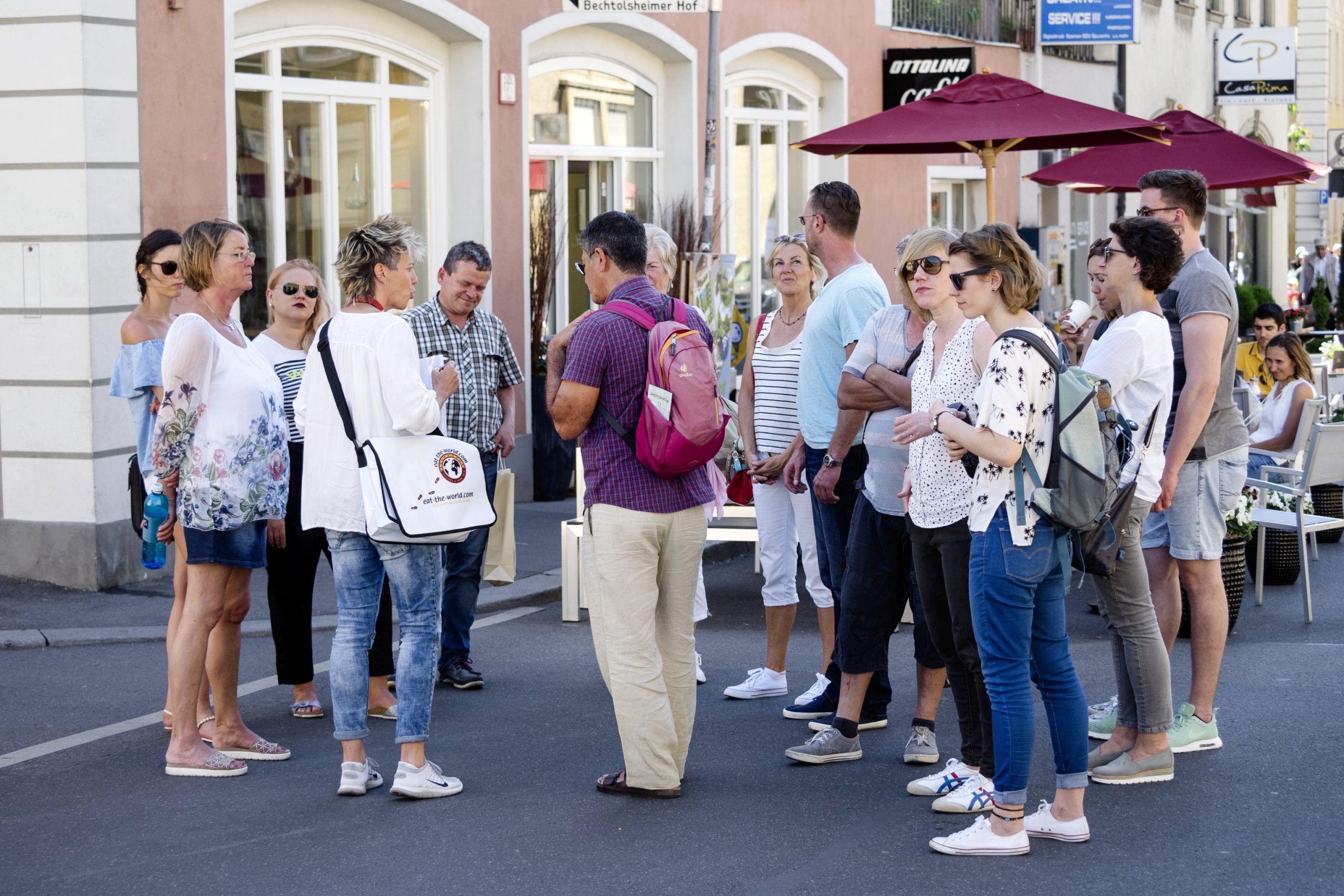 Eine Gruppe von Menschen steht auf einer Straße, manche mit Rucksäcken, plaudert angeregt über ihren Betriebsausflug und genießt die lebhafte Atmosphäre und die Gesellschaft der anderen.