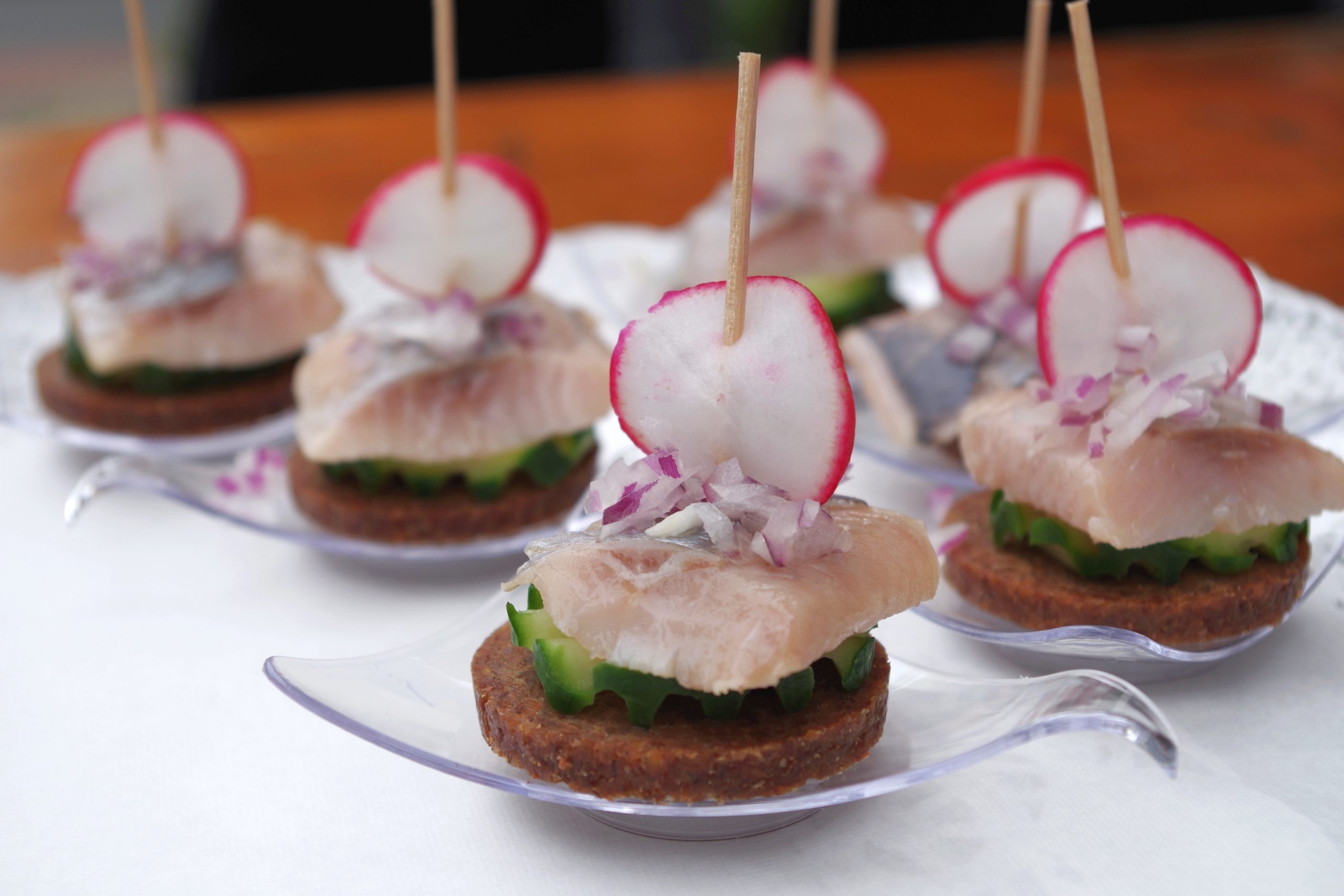 Genießen Sie während des Betriebsausflugs Vorspeisen mit geschnittenem Rettich, Fisch und gewürfelten Zwiebeln auf Brotscheiben, jeweils elegant auf einem kleinen durchsichtigen Löffel serviert.