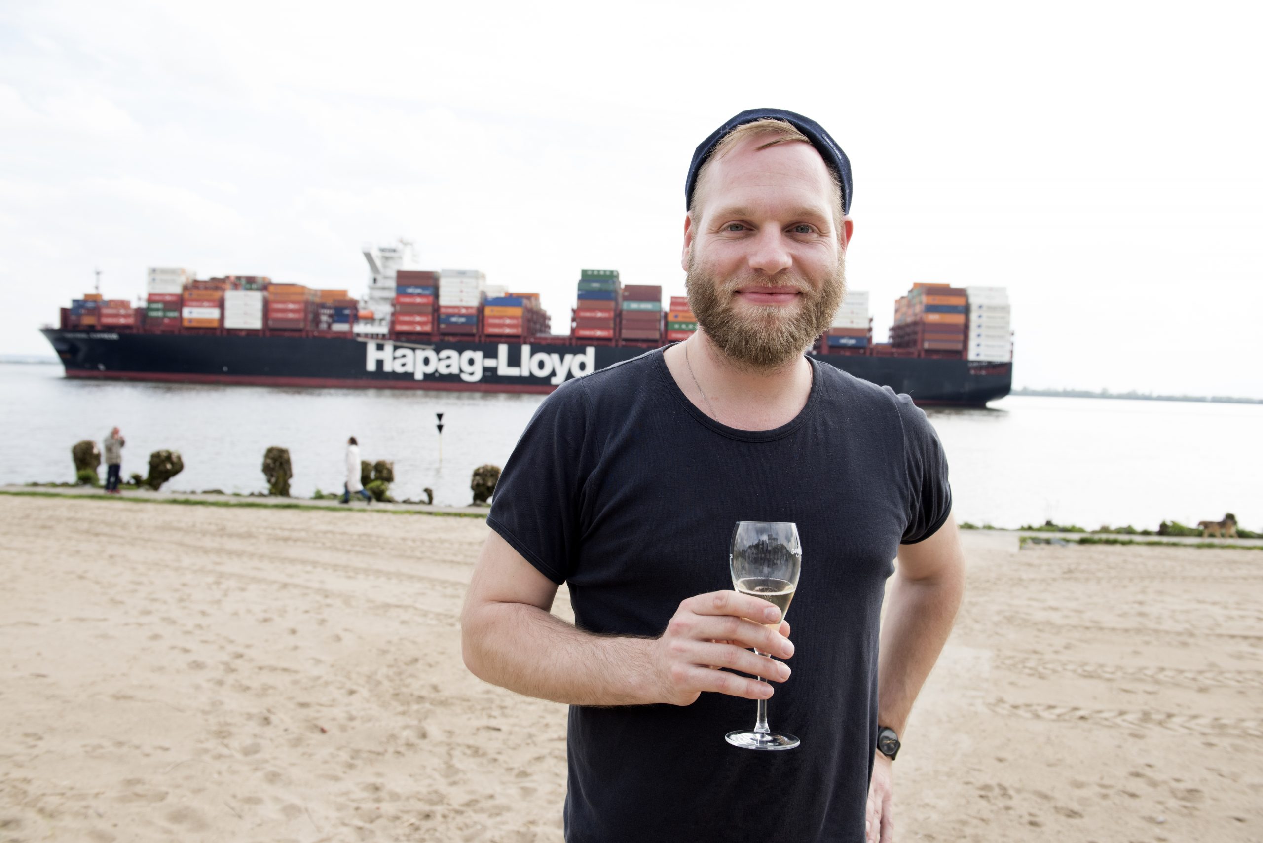 Ein Mann genießt während eines Betriebsausflugs ein Glas Wein an einem Sandstrand, vor der ruhigen Kulisse eines großen Frachtschiffs, das über das Wasser gleitet.