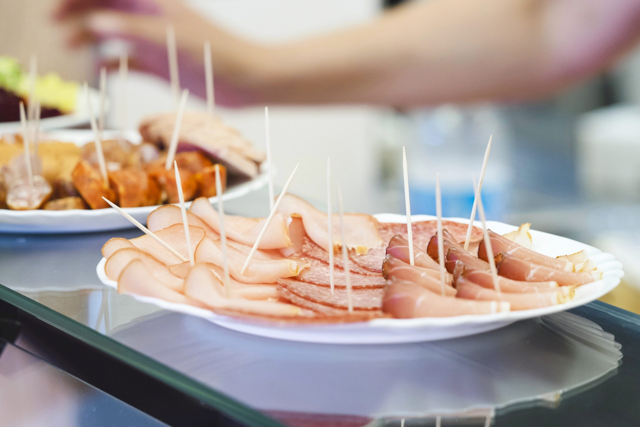 Nahaufnahme von aufgeschnittenem Fleisch auf einem Teller mit Zahnstochern, der auf einer Theke steht. Im Hintergrund streckt sich die Hand einer Person, wahrscheinlich von der Betriebsausflug-Gruppe, herüber.