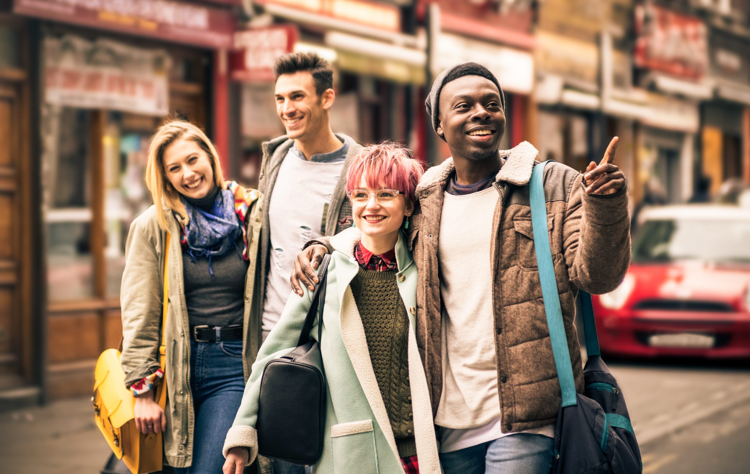 Vier Freunde schlendern während eines Betriebsausflugs die Straße entlang, lächeln und plaudern, während einer von ihnen aufgeregt auf etwas in der Ferne zeigt.