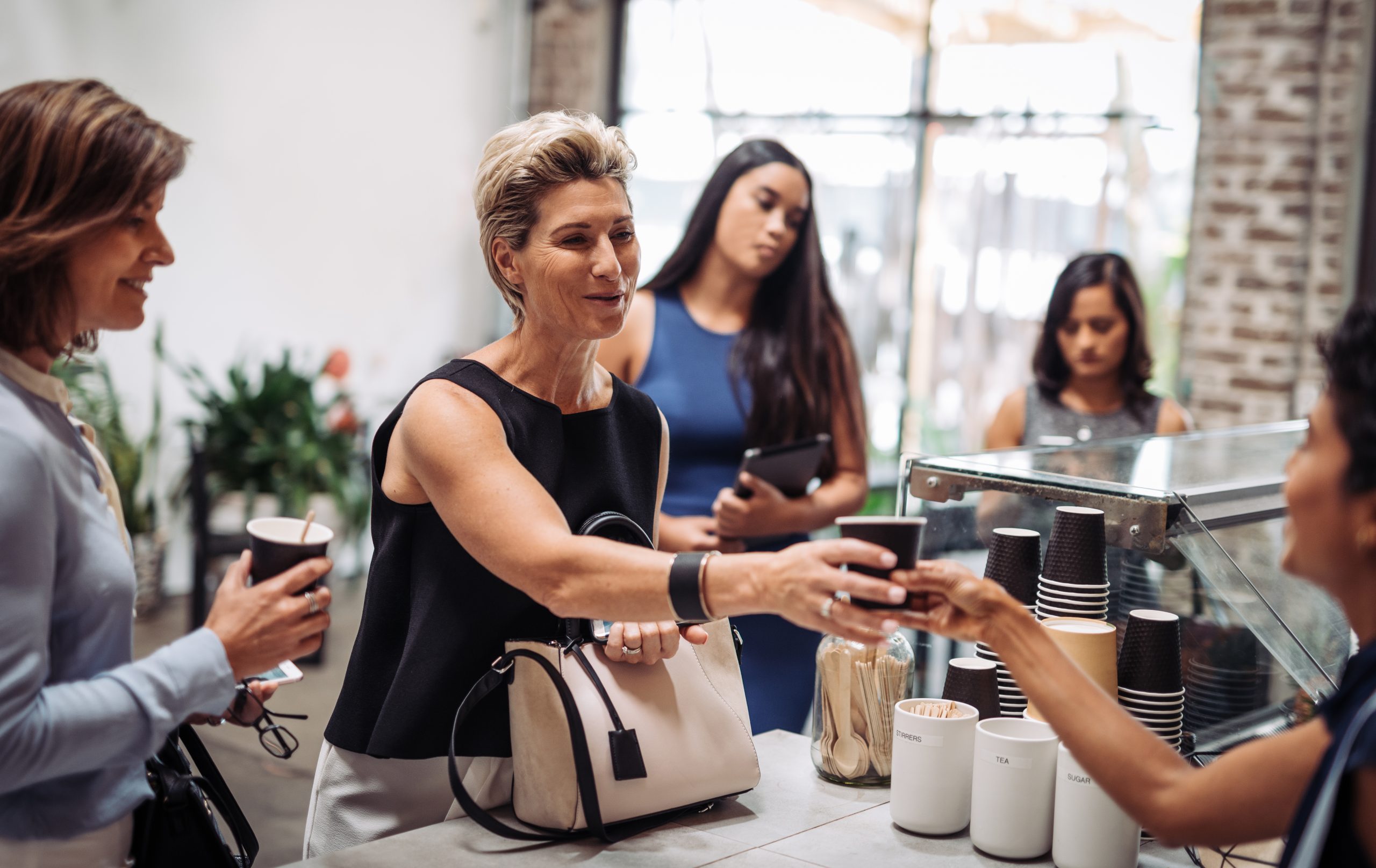 Bei einem lebhaften Betriebsausflug treffen sich Frauen an der Café-Theke, halten ihre dampfenden Tassen Kaffee in der Hand und tauschen angeregte Gespräche aus.