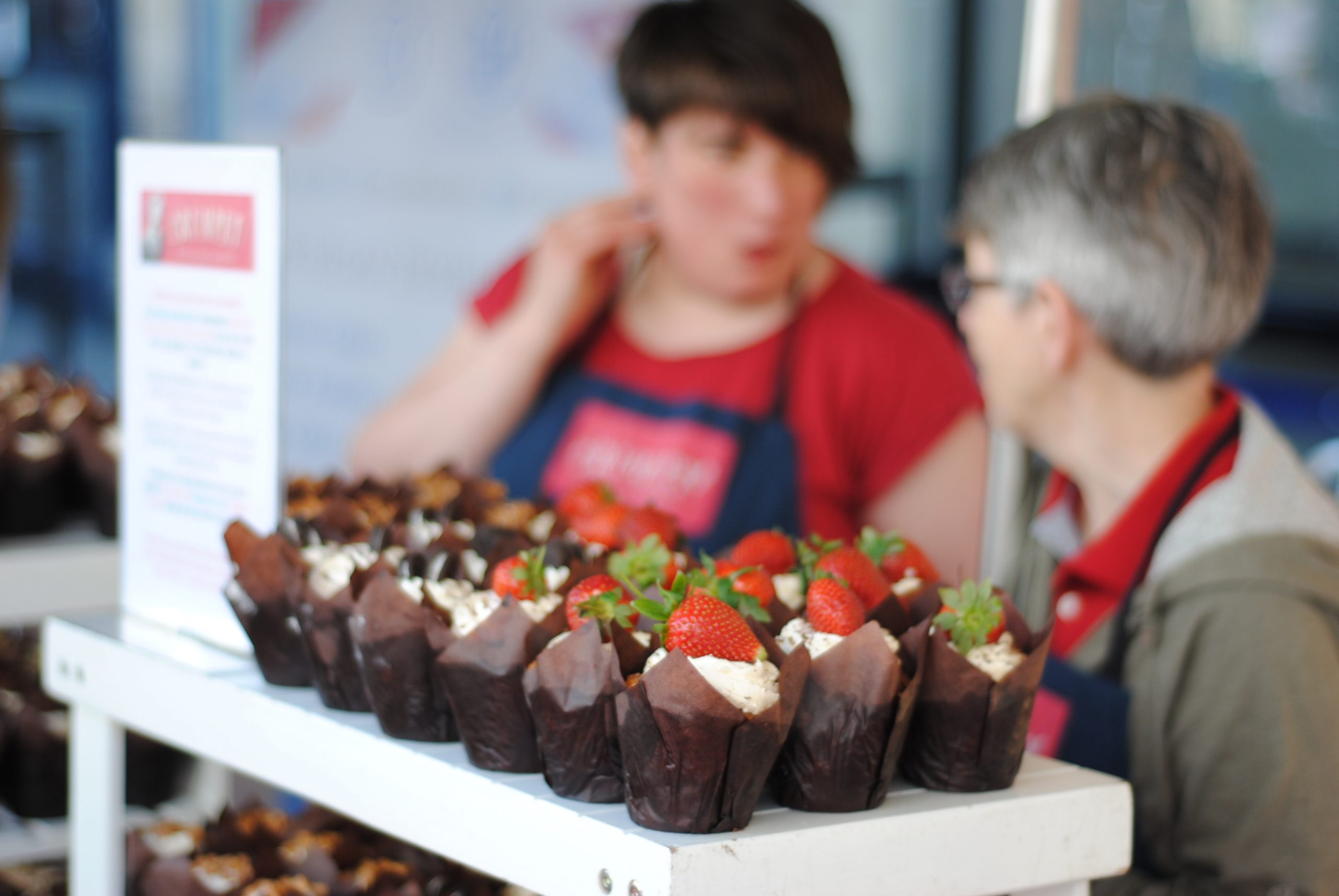 Zwei Kollegen hinter einer Auslage mit Schokoladen-Cupcakes und Erdbeeren an einem Imbissstand während ihres Betriebsausflugs.