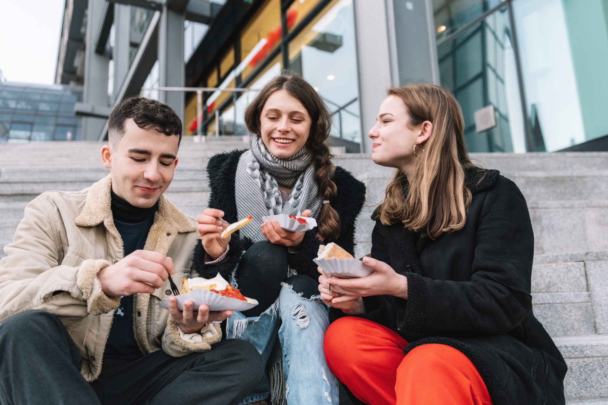 Drei Freunde sitzen auf einer Außentreppe, lächeln und genießen Pommes, die auf Pappschalen serviert werden, und machen offenbar einen vergnüglichen Betriebsausflug.