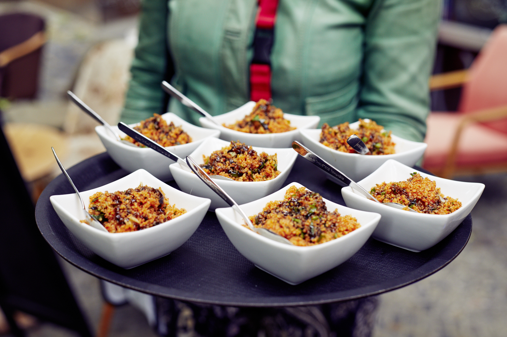 Ein Tablett mit acht quadratischen Schüsseln Quinoa-Salat, jeweils mit einem Löffel, gehalten von einer Person in einer grünen Jacke, perfekt zum Teilen während eines Betriebsausflugs.