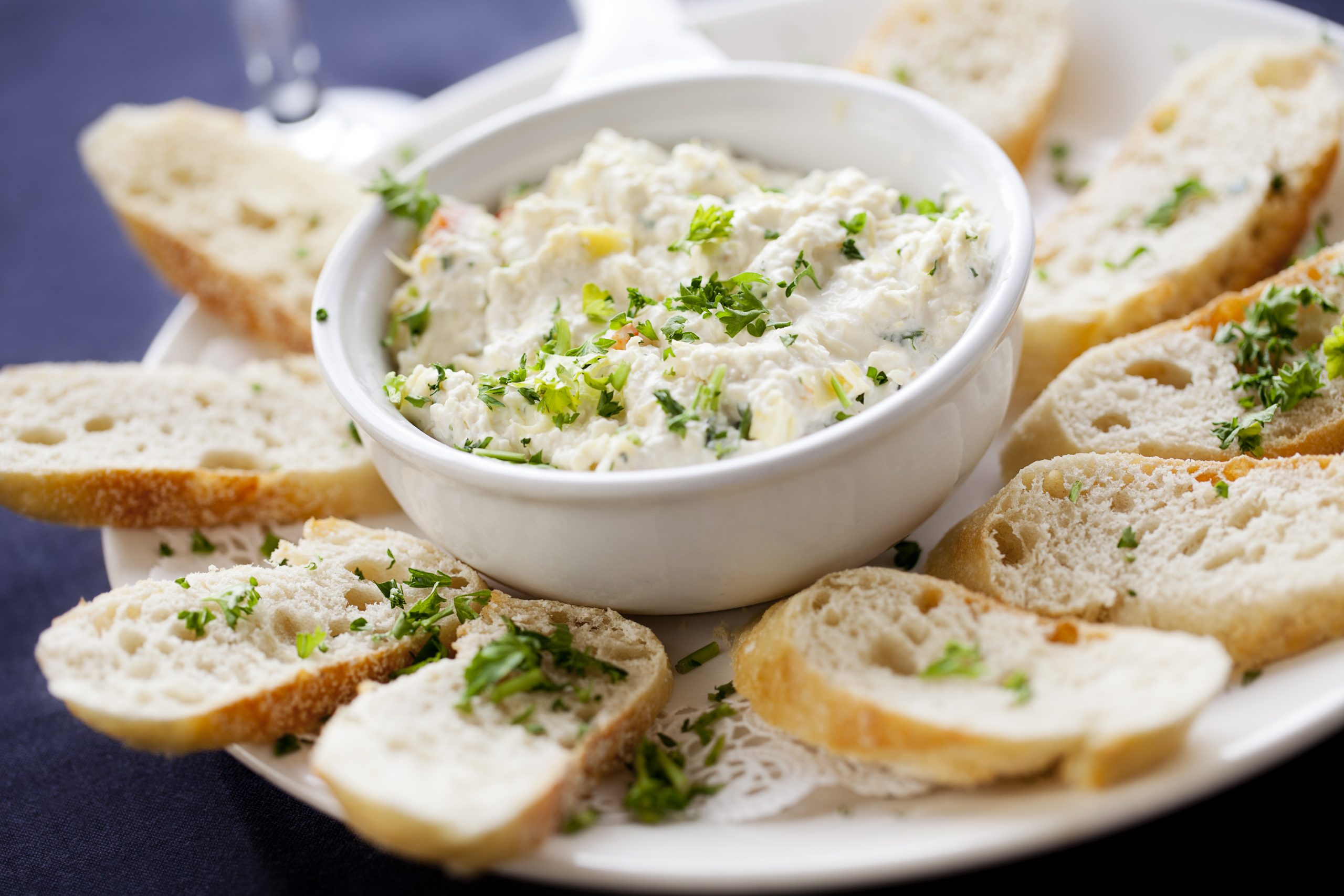 Eine Schüssel cremiger Dip, perfekt für einen Betriebsausflug, garniert mit frischen Kräutern und umgeben von Brotscheiben auf einem weißen Teller.