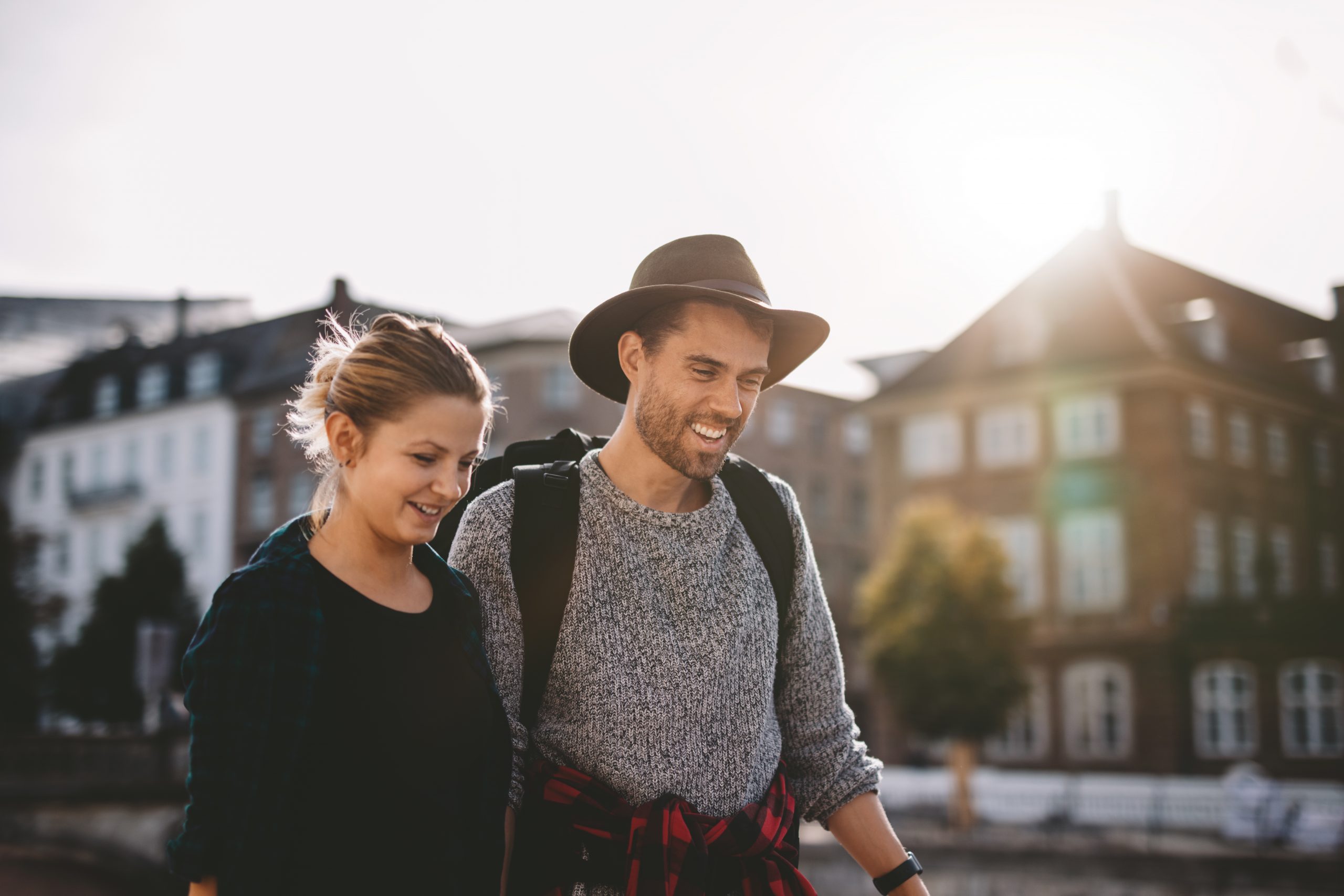 Ein lächelndes Paar genießt einen Betriebsausflug und schlendert gemeinsam im Freien in der Nähe von Gebäuden umher, während im Hintergrund die warm scheinende Sonne scheint.