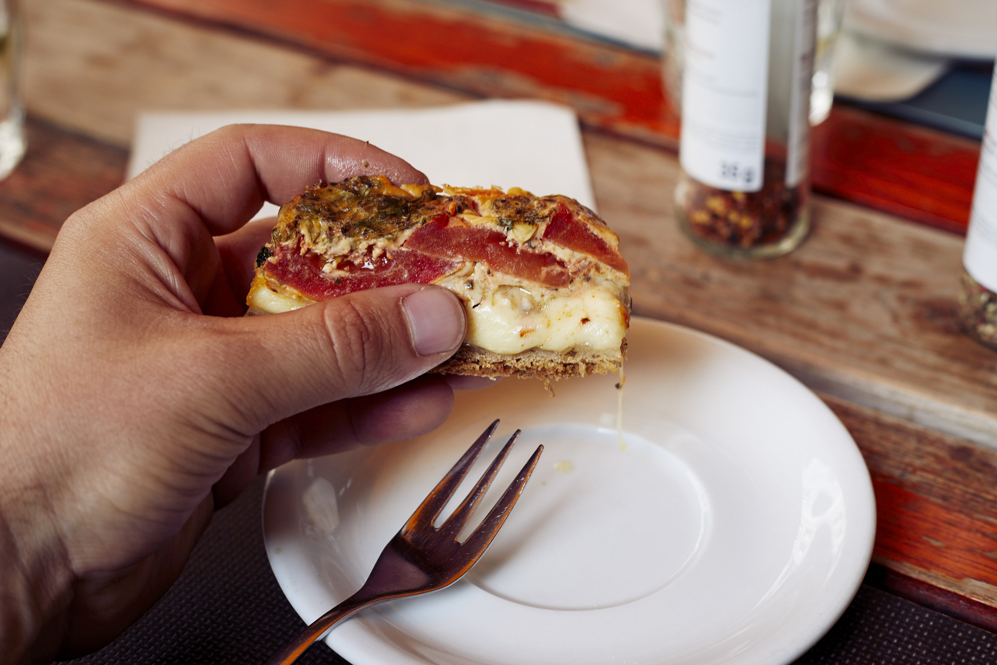 Bei einem Betriebsausflug hält eine Hand ein herzhaftes Sandwich mit Fleisch und Käse, das über einem weißen Teller mit einer Gabel auf dem rustikalen Holztisch balanciert.