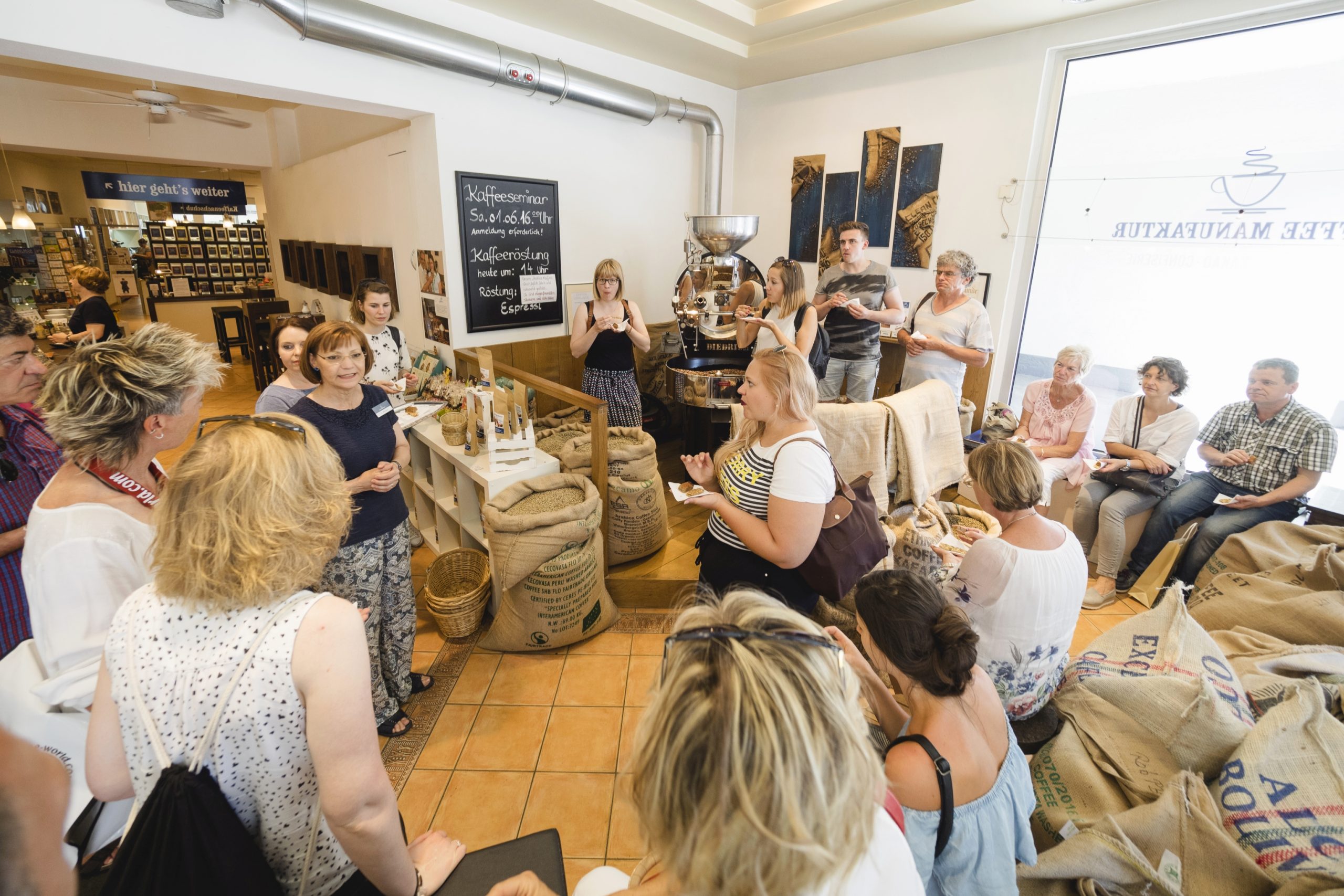 Während eines lebhaften Betriebsausflugs versammelten sich die Kollegen in einem gemütlichen Café und hörten aufmerksam einer Präsentation inmitten einer einladenden Auswahl an Kaffeebeuteln und Geräten zu.