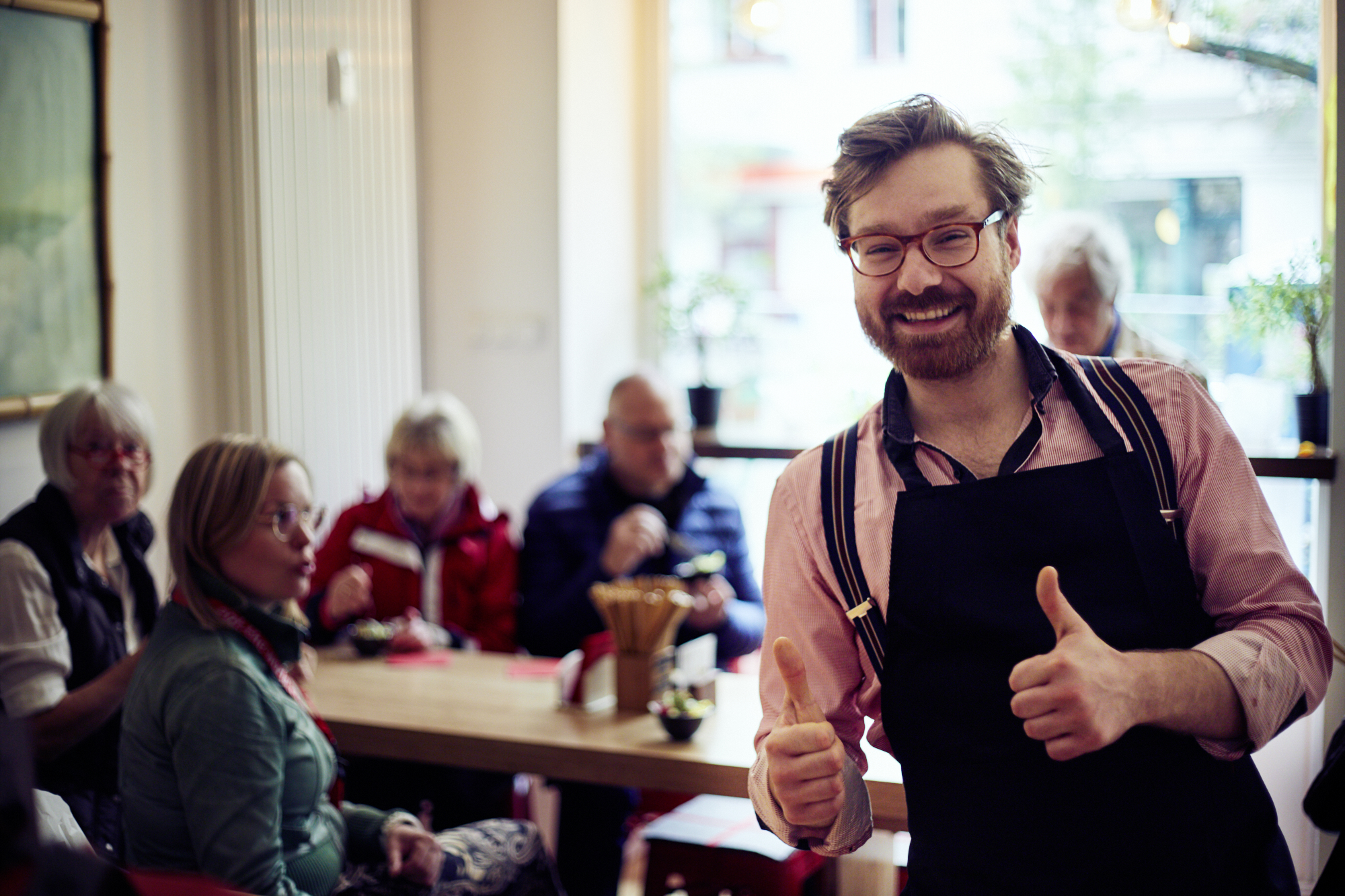 Ein lächelnder Barista zeigt in einem gemütlichen Café mit freundlicher Atmosphäre den Daumen nach oben, perfekt für einen Betriebsausflug, während im Hintergrund die Leute an den Tischen ihre Zeit genießen.