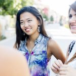 Drei Frauen lächeln und unterhalten sich an einem sonnigen Tag im Freien, mit Bäumen und Gebäuden im Hintergrund.