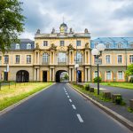 Ein prächtiges Gebäude mit einem Torbogen überspannt eine Straße mit üppigen Bäumen und einem bewölkten Himmel im Hintergrund.