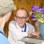 Älterer Mann mit Brille und Schnurrbart lächelt einen Kunden neben einer Theke mit einer Vase mit lila Blumen an.