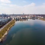 Luftaufnahme einer Stadt mit einem großen See, umgeben von modernen Gebäuden und Grünflächen unter einem teilweise bewölkten Himmel.