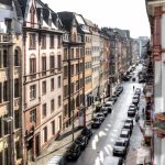 Blick auf eine Stadtstraße gesäumt von historischen europäischen Gebäuden und geparkten Autos an einem sonnigen Tag.