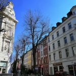 Straßenansicht mit historischen Gebäuden, blattlosen Bäumen und einem strahlend blauen Himmel an einem sonnigen Tag.