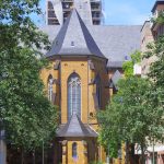 Historische Backsteinkirche mit hohem Turm, umgeben von Bäumen und modernen Gebäuden unter einem klaren blauen Himmel.