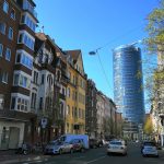 Straßenansicht mit abwechslungsreicher Architektur, geparkten Autos und einem hohen gläsernen Bürogebäude im Hintergrund.