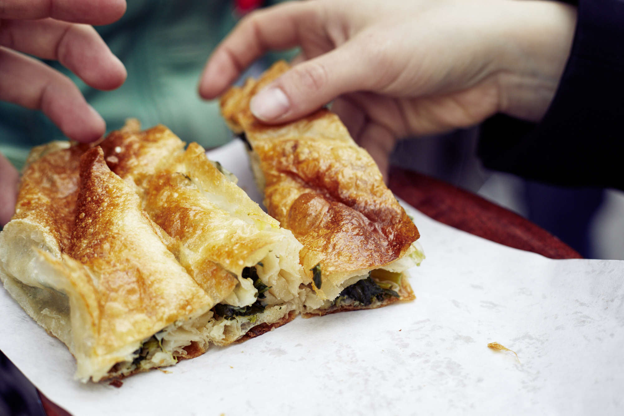 Zwei Hände greifen gierig nach Blätterteigstücken, die mit Spinat und Käse gefüllt sind und auf einem weißen Blatt Papier liegen. Es ist ein alltäglicher Moment, der in Erlebnisgastronomie verwandelt wird und die Essenz eines kulinarischen Abenteuers in einem einfachen, aber köstlichen Snack einfängt.