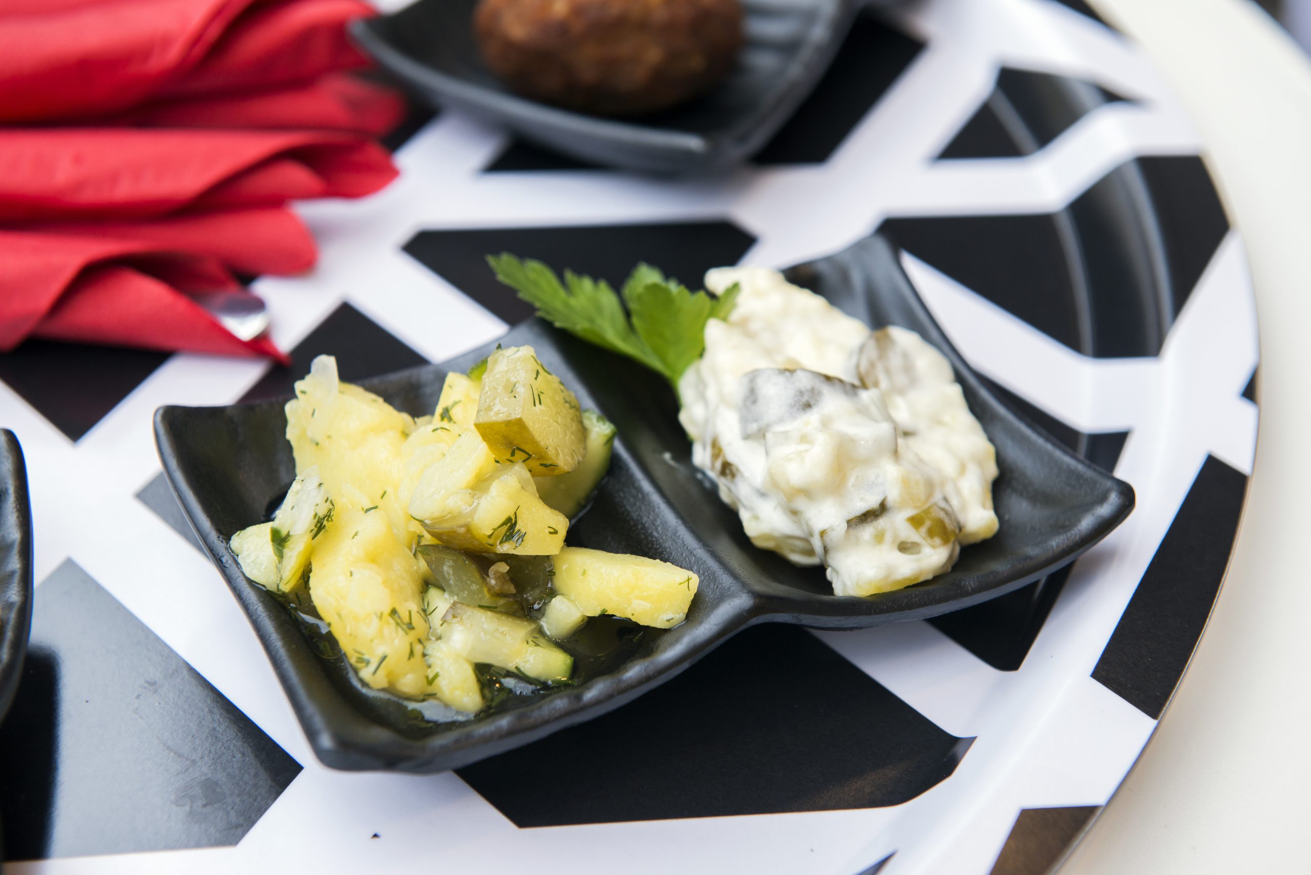 Zwei schwarze Gerichte mit Kartoffelsalat und cremigem Gurkensalat ruhen elegant auf einem Teller mit schwarz-weißem Zickzack-Muster und machen die gesamte Präsentation zu einem wahren Genuss der Erlebnisgastronomie.