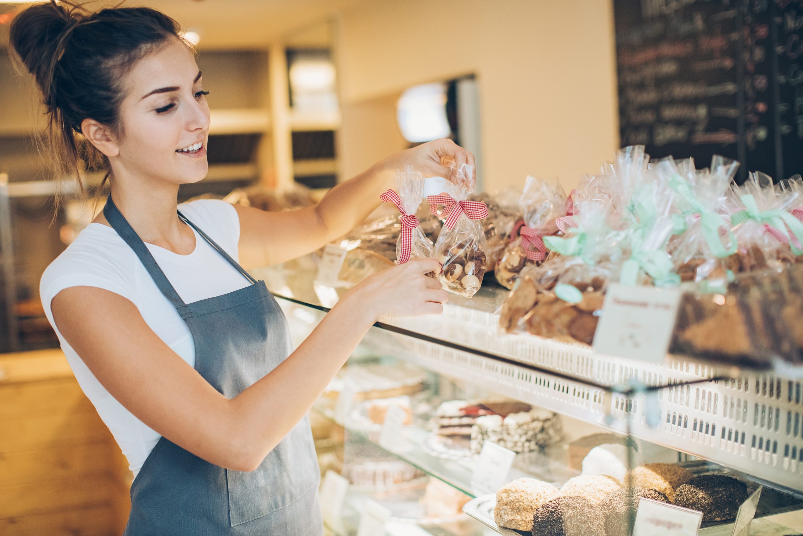 Eine Frau in einer Schürze ordnet verpackte Backwaren sorgfältig in einem Ausstellungsregal an und verleiht der gemütlichen Bäckerei einen Hauch von Erlebnisgastronomie.