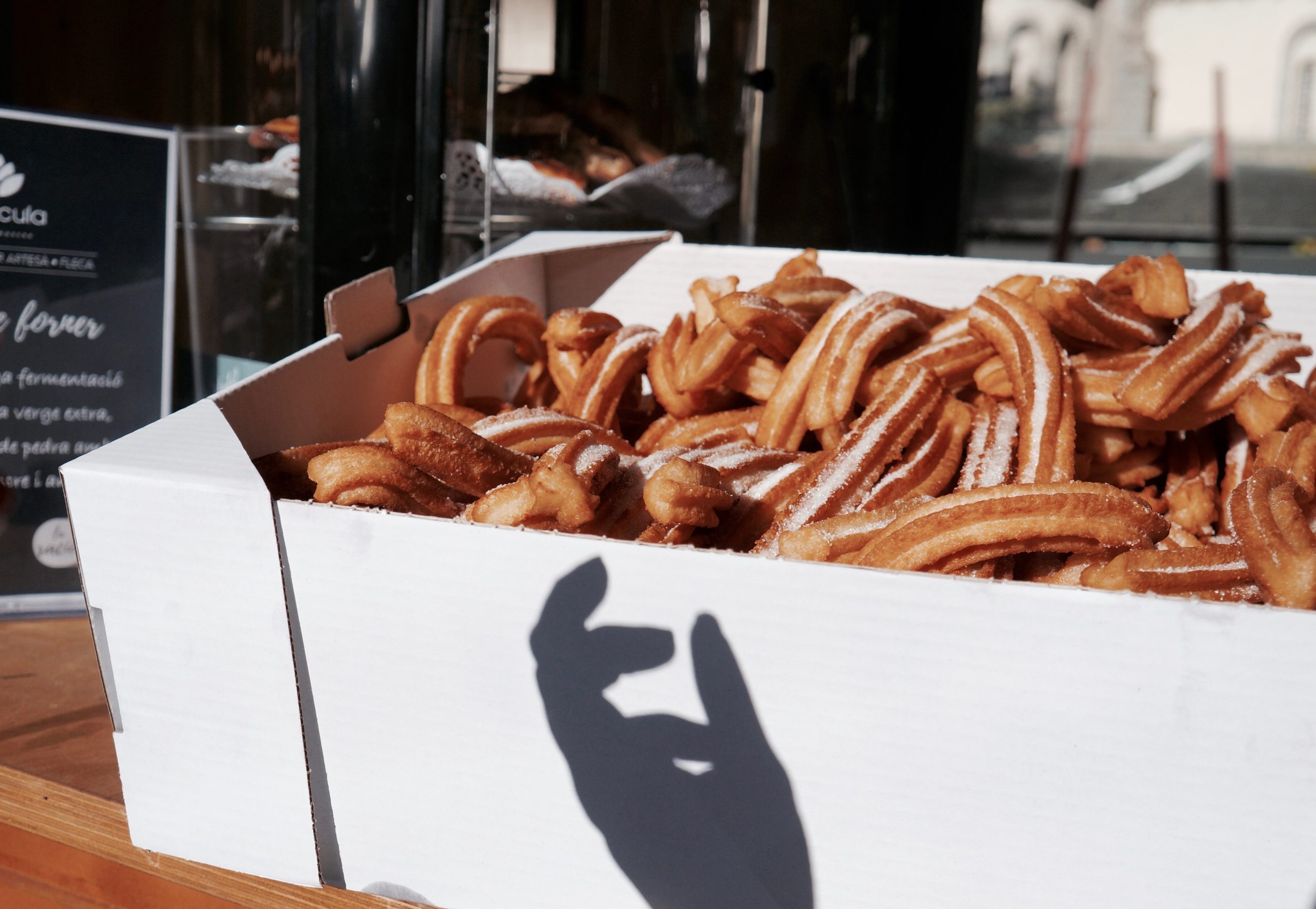 Eine mit Puderzucker bestäubte Schachtel Churros steht daneben, während der Schatten einer Hand eine Geste bildet und der Szene einen Hauch von Erlebnisgastronomie verleiht.