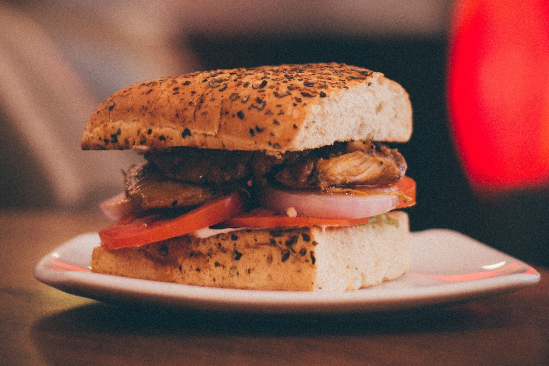 Ein genussvolles Erlebnis in der Erlebnisgastronomie: eine Nahaufnahme eines Sandwiches mit gegrilltem Hähnchen, Tomaten und Zwiebeln auf einem Brötchen mit Körnern, serviert auf einem weißen Teller.