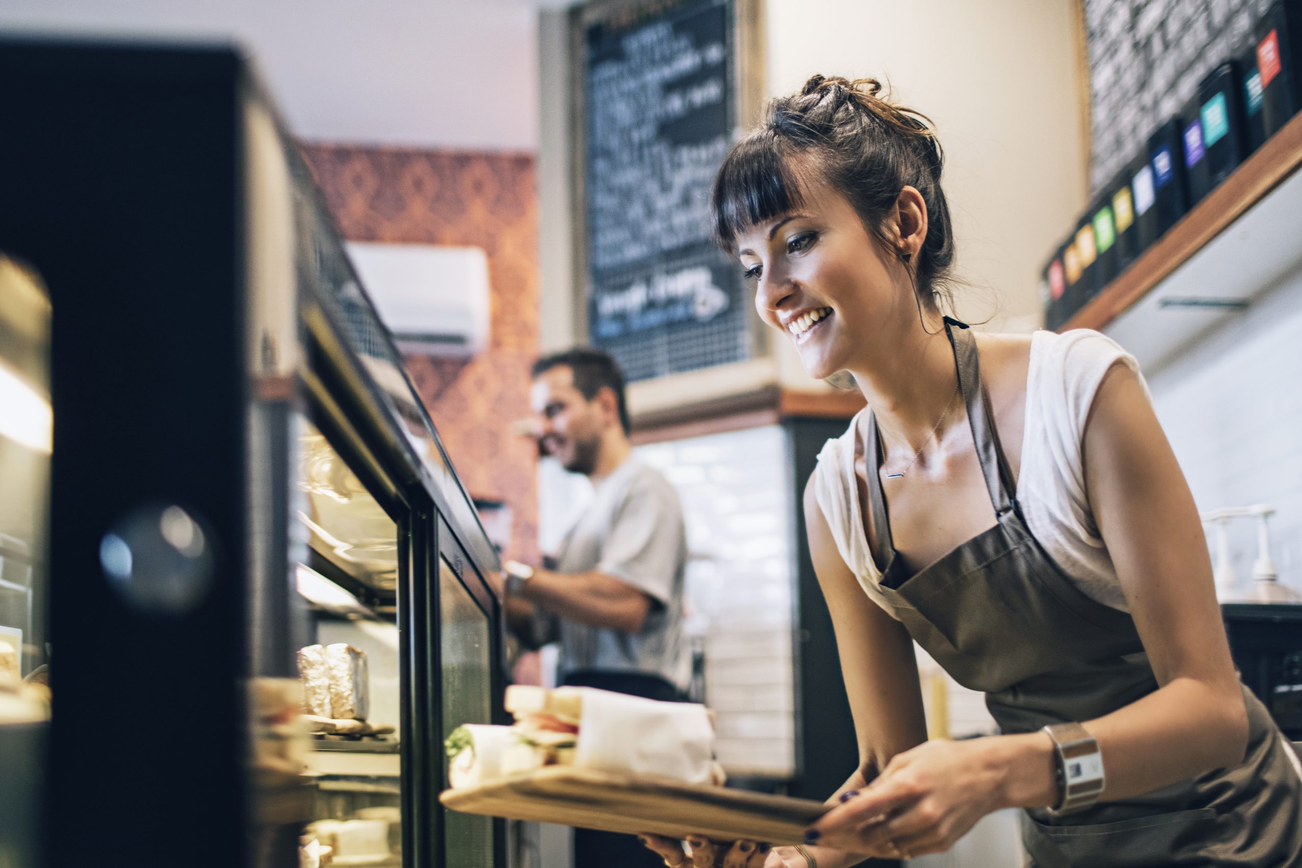 Ein lächelnder Barista in einer Schürze serviert Speisen aus einer Vitrine und bietet einen Hauch von Erlebnisgastronomie in der einladenden Atmosphäre des gemütlichen Cafés.