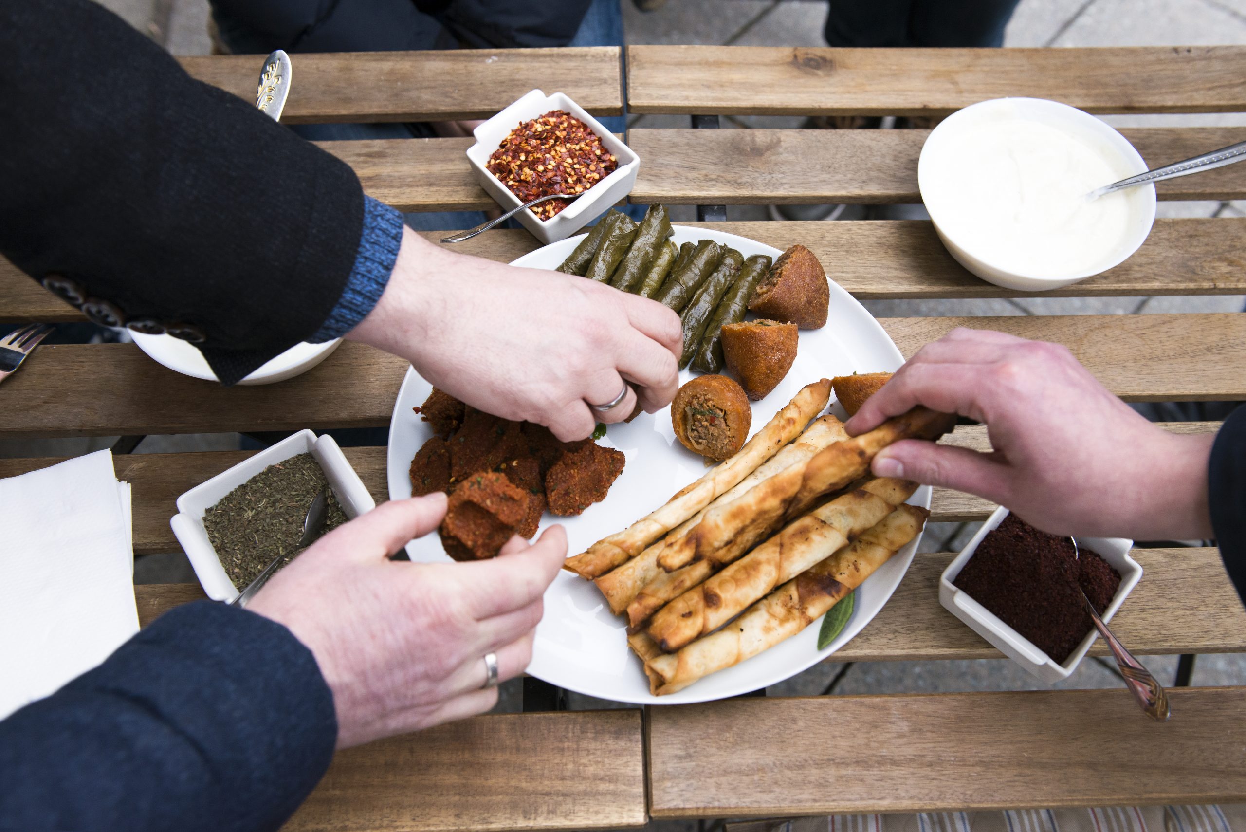 Hände greifen gierig nach verschiedenen Vorspeisen auf einem rustikalen Holztisch und erleben die Essenz der Erlebnisgastronomie mit Grissini, Dips und gefüllten Weinblättern.