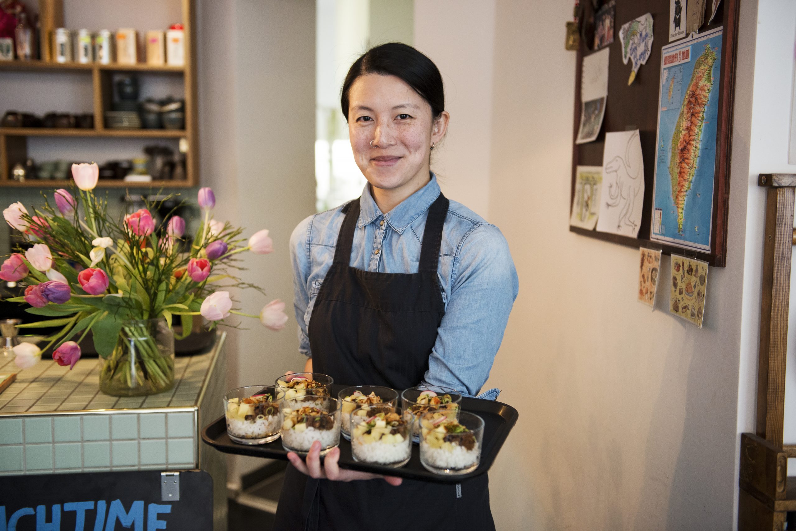 Eine Person in einer Schürze hält ein Tablett mit köstlichen Desserts und verkörpert den Geist der Erlebnisgastronomie in einem gemütlichen, mit wunderschönen Blumen geschmückten Café.