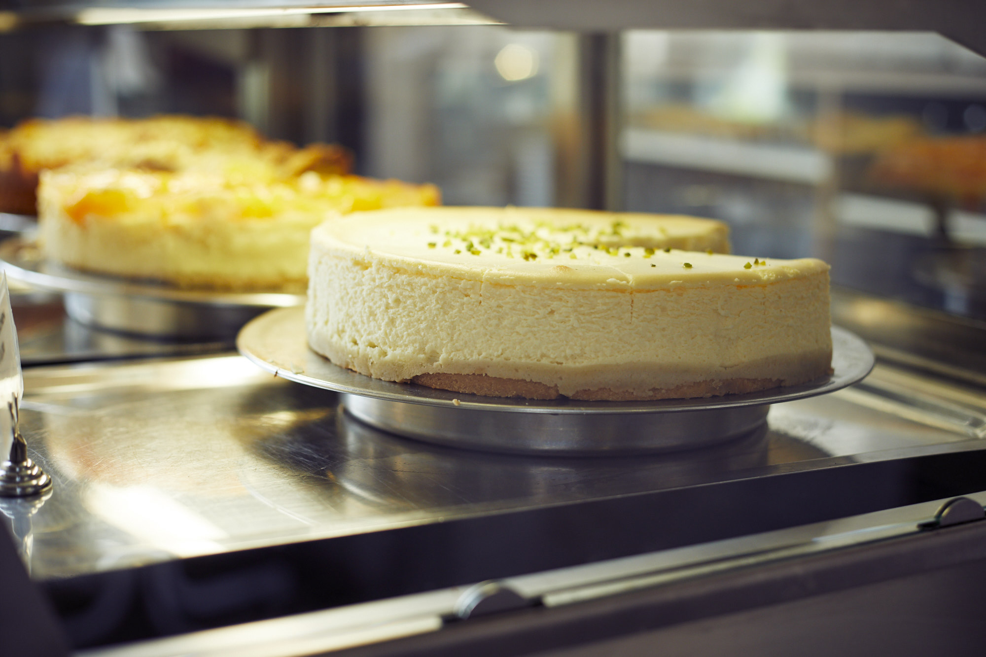 In einer Vitrine der Erlebnisgastronomie steht ein Käsekuchen elegant in einer Glasvitrine, von dem ein Stück fehlt, und seine cremige Oberfläche ist mit leuchtend grünen Streuseln verziert.